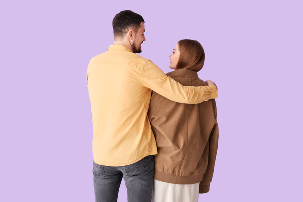 Joven abrazando esposa en su chaqueta sobre fondo lila, vista trasera - Foto, imagen