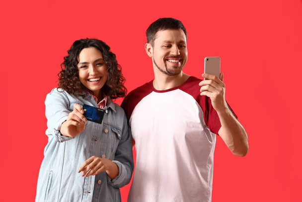 Junges Paar mit Kreditkarte und Handy auf rotem Hintergrund - Foto, Bild