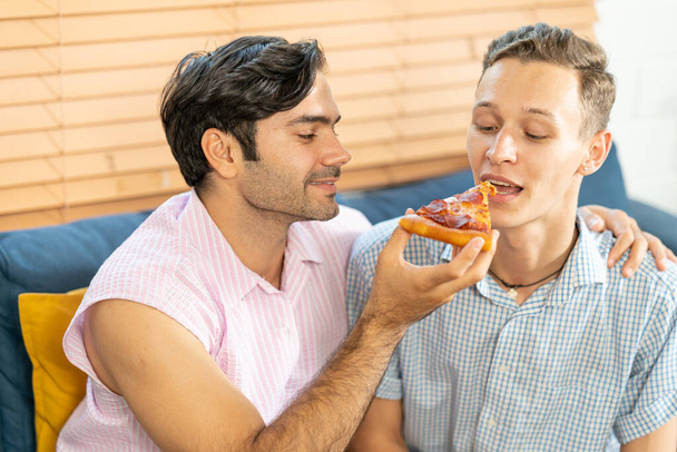 Cheerful gay couple eating pizza. Gay couple sitting on sofa at home,  eating one slice of pizza. Homosexual relationship concept. - Foto, afbeelding