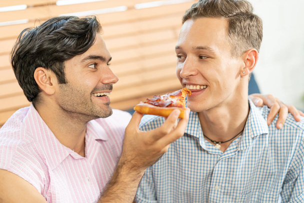 陽気なゲイのカップルはピザを食べる。ゲイのカップル自宅でソファに座って、ピザの1つのスライスを食べる。同性愛関係の概念. - 写真・画像
