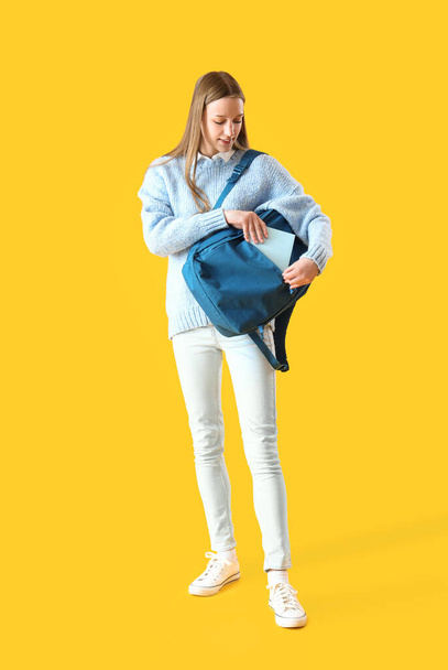 Female student putting copybook into backpack on yellow background - 写真・画像