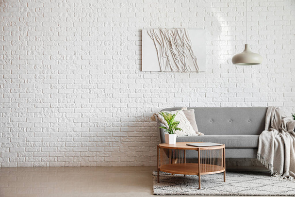 Szara sofa i stolik kawowy z nowoczesnym laptopem w pobliżu ściany z białej cegły - Zdjęcie, obraz