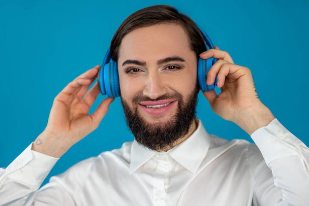 Happy cheerful transgender man listening his favorite songs on headphones looking at camera with smile and joyful face isolated over blue background - Foto, Imagen
