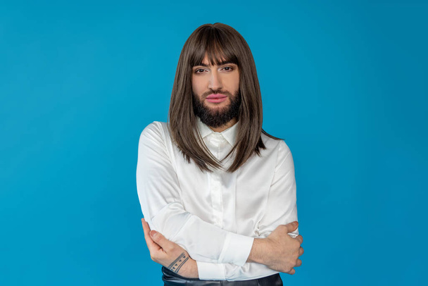 Attractive transgender person wearing brunette wig and white shirt hugging himself protects personal individuality posing isolated over blue background - Valokuva, kuva
