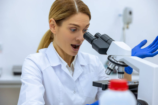 Amazed medical female scientist looking under microscope, doing analysis of test sample, ambitious biotechnology specialist working in laboratory. - Foto, Imagem