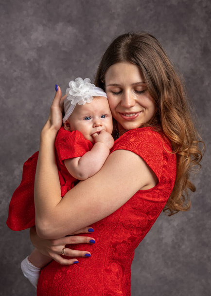 Mama knuffelt zachtjes haar dochtertje. Gekleed in rode jurken. Grijze achtergrond.Studio schieten. De liefde van moeder. Pasgeboren zorg. Gezinsconcept - Foto, afbeelding