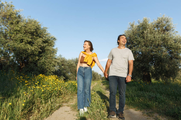 Снимают влюблённую молодую пару, идущую по травяному полю. Мужчина и женщина ходят по высокой траве и оливковым деревьям - Фото, изображение