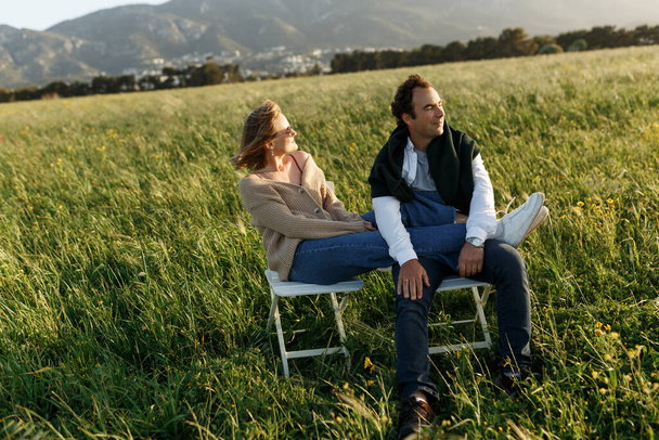 若いです幸せなカップルに座って椅子に羽の草のフィールドで夏の日没の日. - 写真・画像