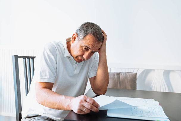 Middle aged man holding documents reading paper bills, calculating pension fees, payments, taxes, planning family retirement money finances at home. middle-aged man at table at home reading bill - Foto, afbeelding