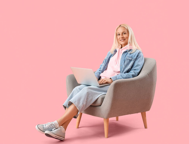 Mature female programmer working with laptop in armchair on pink background - Фото, зображення