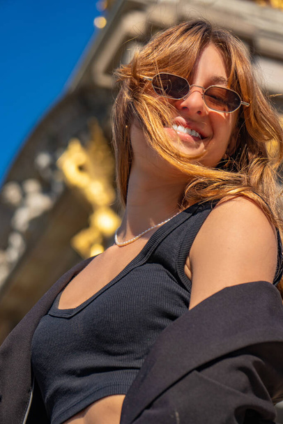 Beautiful young model with blonde hair blowing in the wind, wearing sunglasses and a black top laughing and smiling happily with a beautiful sunlit face and a black jacket falling from her shoulders - Foto, Bild