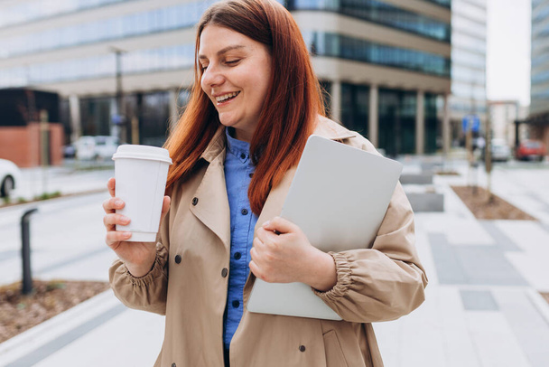 Caucasian redhead businesswoman in fashion office clothes posing outside with a laptop and cup of coffee or tea. Business on the go concept. - Fotó, kép