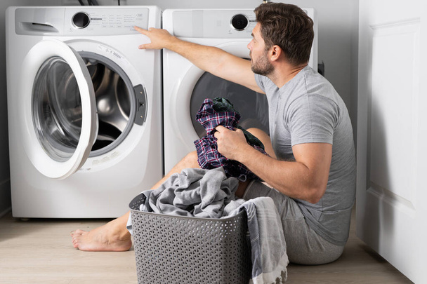 Middle aged single man with laundry basket and dirty clothes near washing machine at home. Millennial man putting clothes into washing machine. Displeased hispanic man wash clothes in washing machine - Fotografie, Obrázek