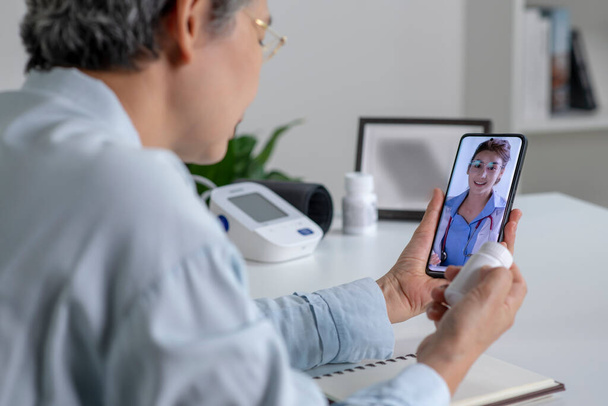 Donna asiatica con smartphone durante una consultazione online con il suo medico nel suo salotto, concetto di telemedicina - Foto, immagini