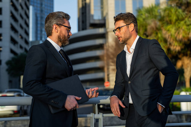 Business communication. Two business men talking outdoors. Two diverse businessmen talking together. Colleagues discussing and thinking about project. Walking and talking between two business men - Foto, afbeelding