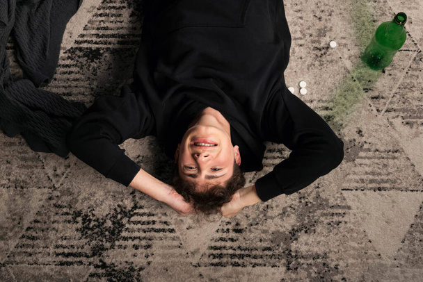 A severely depressed guy, lying on the floor among pills and a bottle of alcohol. A young man in a hard mental condition. alcohol and drugs addiction. Top view. view from above. - Photo, Image