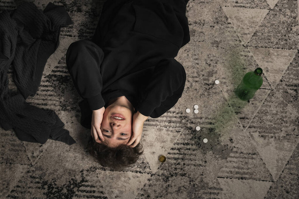 Un gars gravement déprimé, allongé sur le sol parmi des pilules et une bouteille d'alcool. Un jeune homme dans un état mental difficile. alcoolisme et toxicomanie. Vue de dessus. vue d'en haut. - Photo, image
