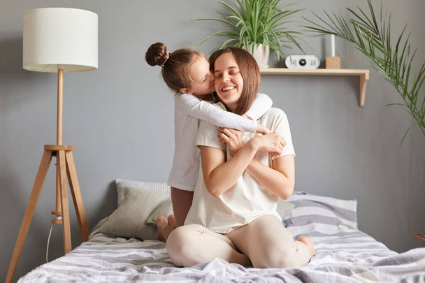 Cute charming kid kissing and hugging mother, daughter with mom sitting in bed in bedroom enjoying time together child congratulating with mother's day. - Fotografie, Obrázek