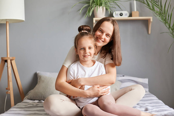 Home portrait of smiling woman and little female child mother and daughter sitting in bed hugging in bedroom looking at camera expressing happiness, mother's day congratulating. - Photo, Image
