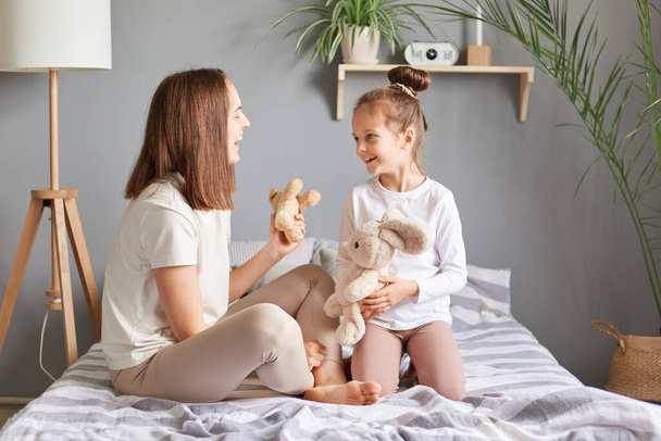 Side view portrait of smiling mother and daughter sitting in bed playing soft toys talking having fun communicating enjoying happy time, childhood, parenthood. - Fotografie, Obrázek