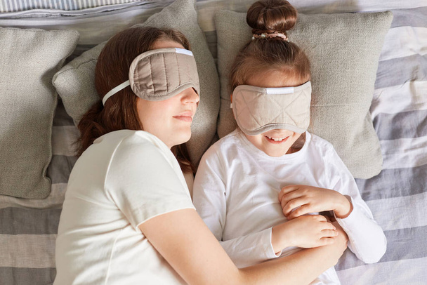 Top view of mother lying on pillow with daughter, wearing blindfolds, resting in morning in bedroom, sleeping hugging in bed, lazy weekend. - Fotó, kép