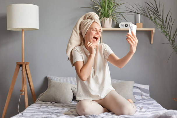 Attractive happy joyful positive cheerful woman wrapped in towel siting in her bed and taking selfie, holding smartphone in hands, having video call on mobile phone. - Фото, зображення