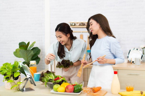 İki genç Asyalı kadın mutfakta bir tablet kullanıyor. Kadın sağlıklı yemek salatasıyla eğleniyor.. - Fotoğraf, Görsel