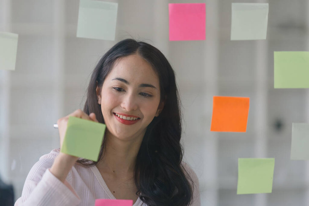 Il concetto di brainstorming è esemplificato come una donna d'affari asiatica millenaria condivide idee usando appunti post-it, che pone su una parete di vetro. - Foto, immagini