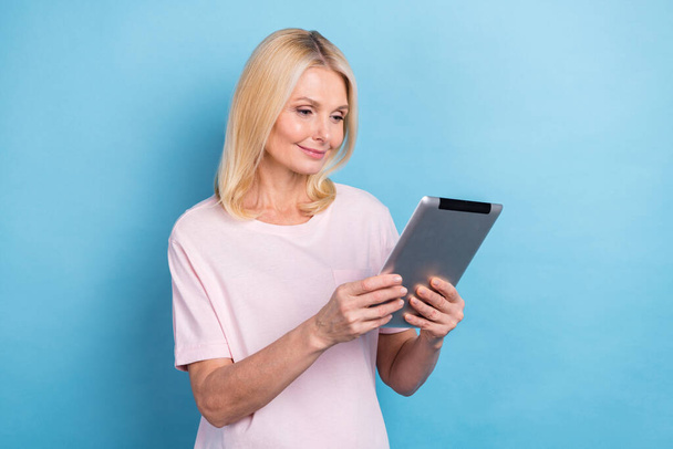 Portrait of good mood pensioner woman with straight hairdo wear white t-shirt read email on tablet isolated on blue color background. - Φωτογραφία, εικόνα