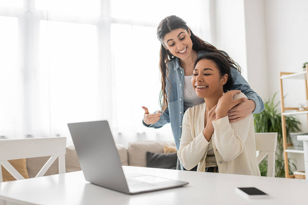χαρούμενη λεσβία γυναίκα που δείχνει την πολυφυλετική φίλη της κοντά στο laptop κατά τη διάρκεια βιντεοκλήσης  - Φωτογραφία, εικόνα