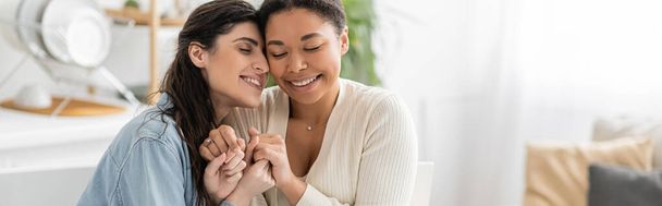 満足している異人種間のレズビアンカップル目を閉じて手を握って自宅、バナー  - 写真・画像