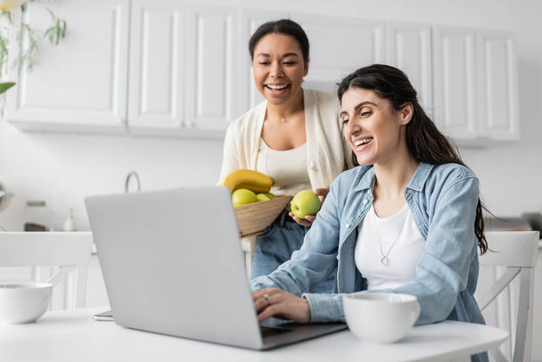 heureuse femme multiraciale tenant bol avec des fruits près de petite amie gaie travaillant sur ordinateur portable à la maison  - Photo, image