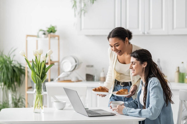 happy multiracial woman holding plates with croissants near girlfriend working on laptop from home  - Photo, Image