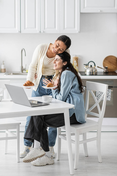 счастливая многорасовая женщина держит тарелки с круассанами рядом с подругой работает на ноутбуке из дома  - Фото, изображение
