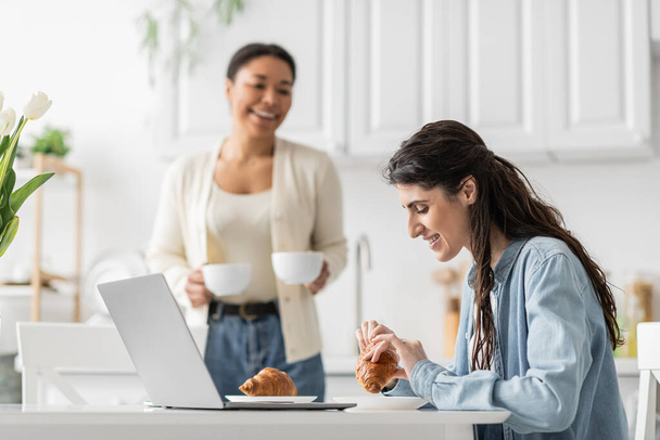 happy lesbian woman eating croissant near laptop and multiracial girlfriend with cups on blurred background  - Foto, Imagen