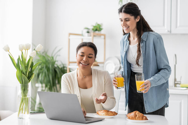 cheerful lesbian woman holding glasses with orange juice near multiracial girlfriend working on laptop  - Photo, Image