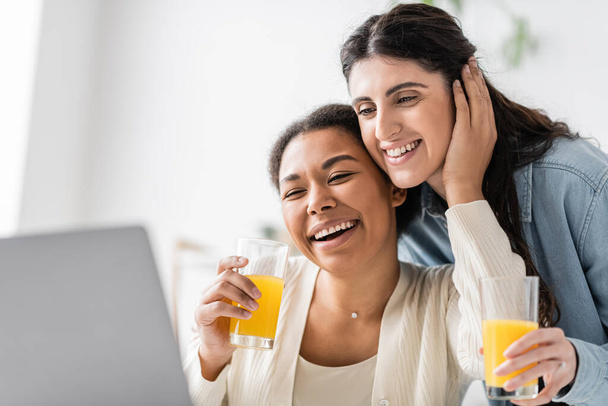 wesoła wielorasowa lesbijska para patrząc na laptopa trzymając szklanki soku pomarańczowego  - Zdjęcie, obraz