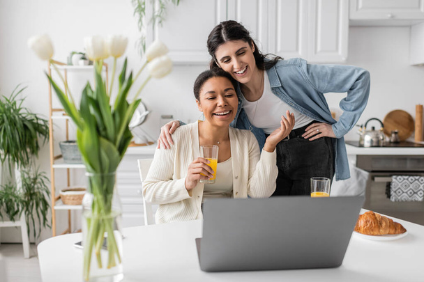 cheerful multiracial lesbian couple looking at laptop while having breakfast in kitchen  - Foto, imagen