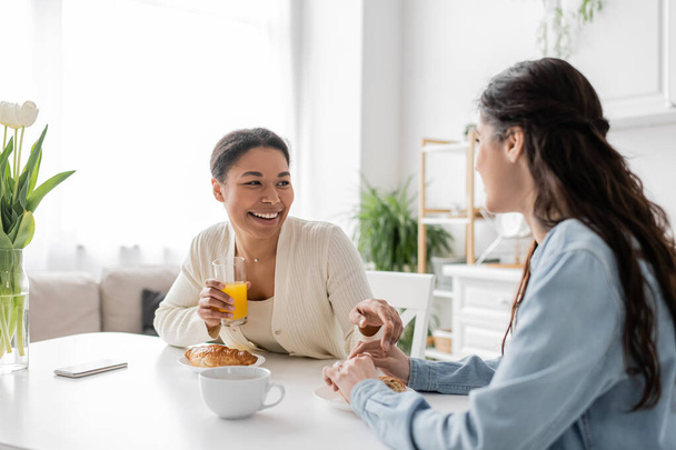 cheerful multiracial lesbian couple smiling while having breakfast in kitchen  - Foto, afbeelding