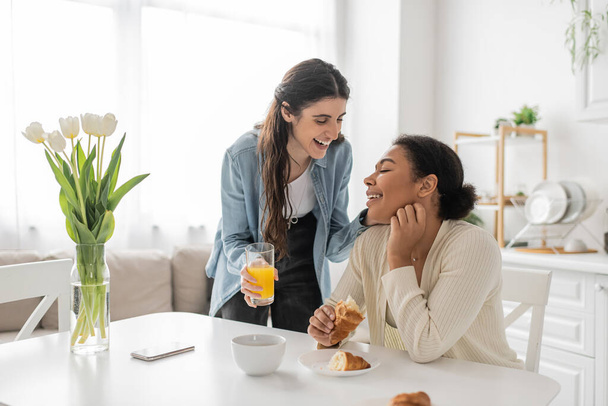 happy lesbian woman holding glass of orange juice near multiracial girlfriend in kitchen  - Fotó, kép