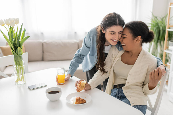 happy lesbian woman holding glass of orange juice and hugging multiracial girlfriend during breakfast in kitchen  - Фото, изображение