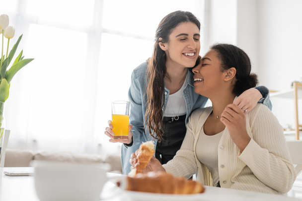 cheerful lesbian woman holding glass of orange juice and hugging multiracial girlfriend during breakfast in kitchen  - Foto, Imagem