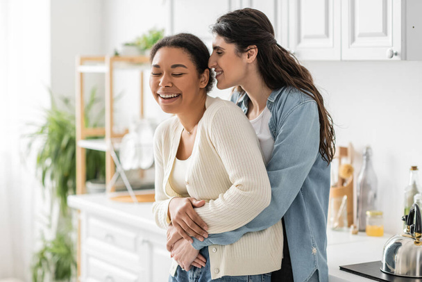 positive lesbian woman hugging multiracial girlfriend in modern kitchen  - Фото, зображення