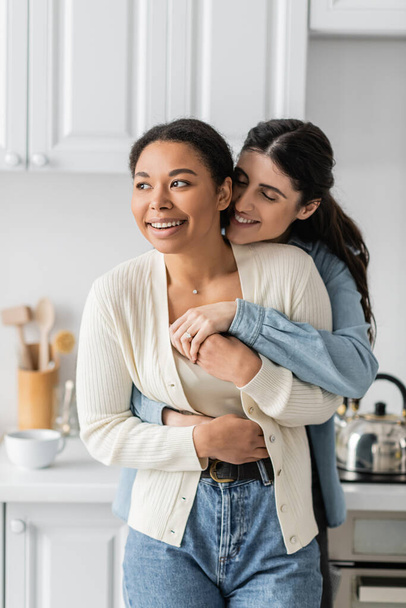 cheerful lesbian woman hugging positive multiracial girlfriend in modern kitchen  - Foto, afbeelding