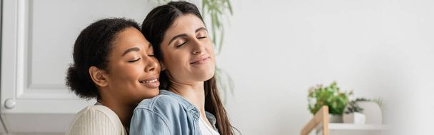 cheerful multiracial lesbian woman with curly hair hugging girlfriend at home, banner  - Fotografie, Obrázek