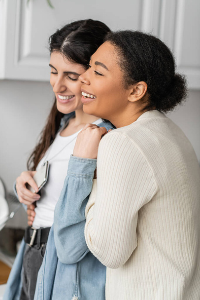 overjoyed lesbian woman holding smartphone and hugging with happy multiracial girlfriend   - Foto, Imagem