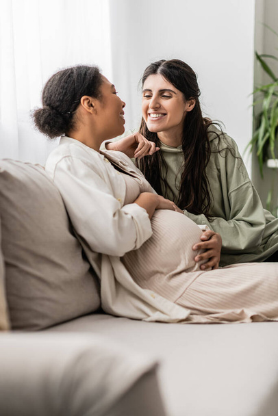 happy lesbian woman hugging pregnant multiracial wife while sitting on couch  - Fotoğraf, Görsel