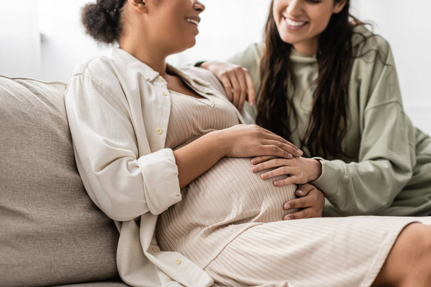 cropped view of happy lesbian woman hugging pregnant multiracial wife while sitting on sofa  - Фото, зображення