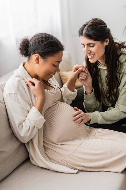 cheerful lesbian woman touching belly of pregnant multiracial wife while sitting on sofa  - Fotoğraf, Görsel