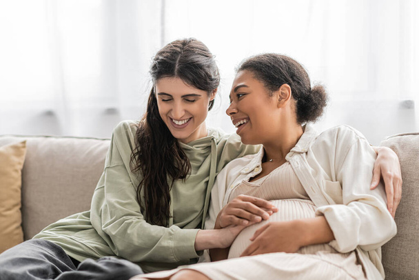 overjoyed lesbian woman hugging pregnant multiracial wife while sitting on sofa  - Foto, imagen